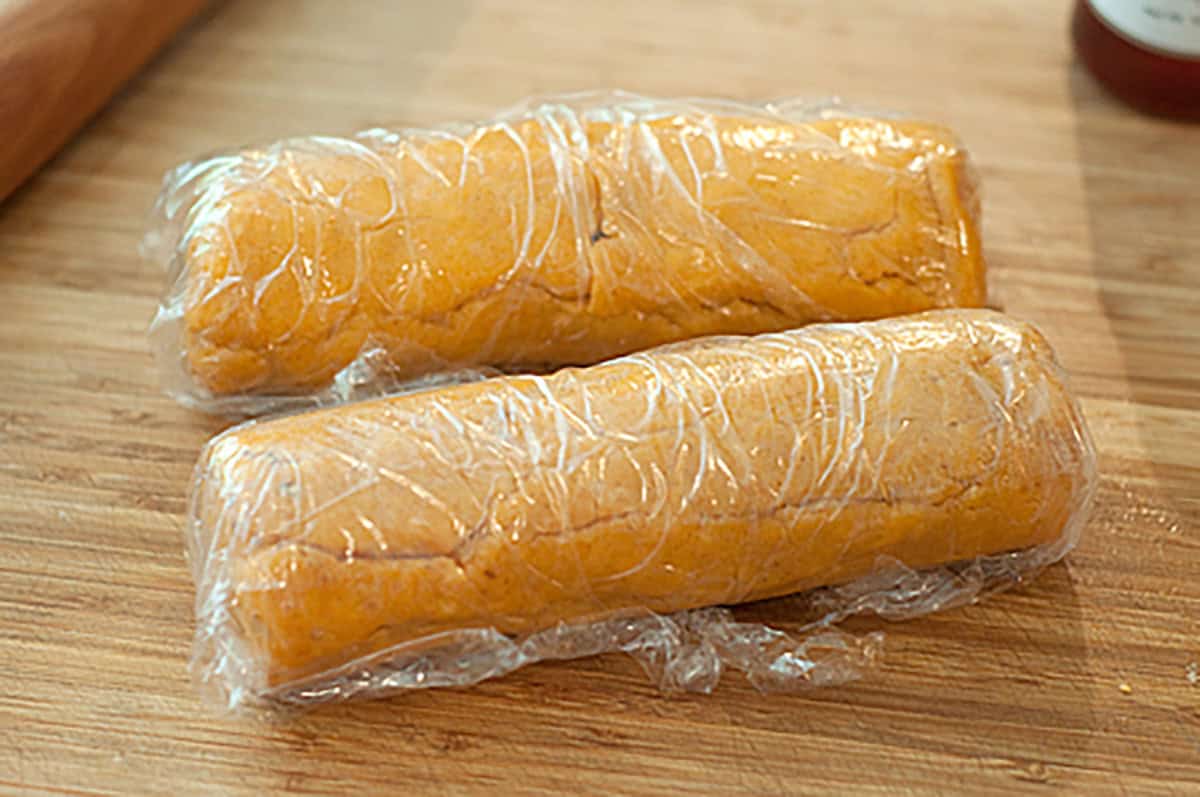 Two logs of yellow dough wrapped in plastic wrap are placed on a wooden surface.
