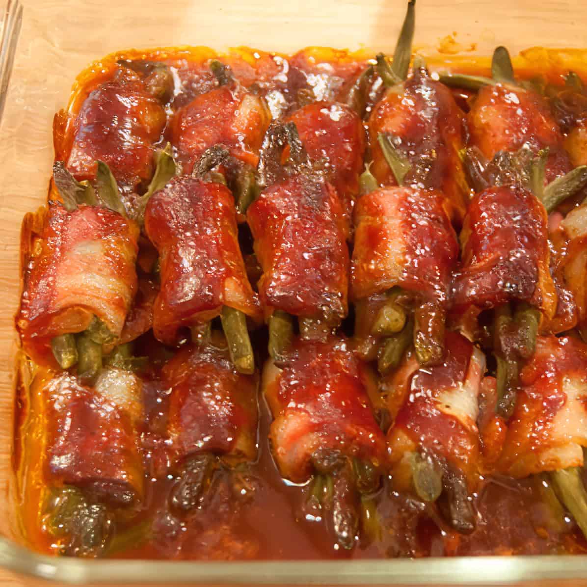 Bacon-wrapped green bean bundles in sauce, arranged in a glass baking dish.