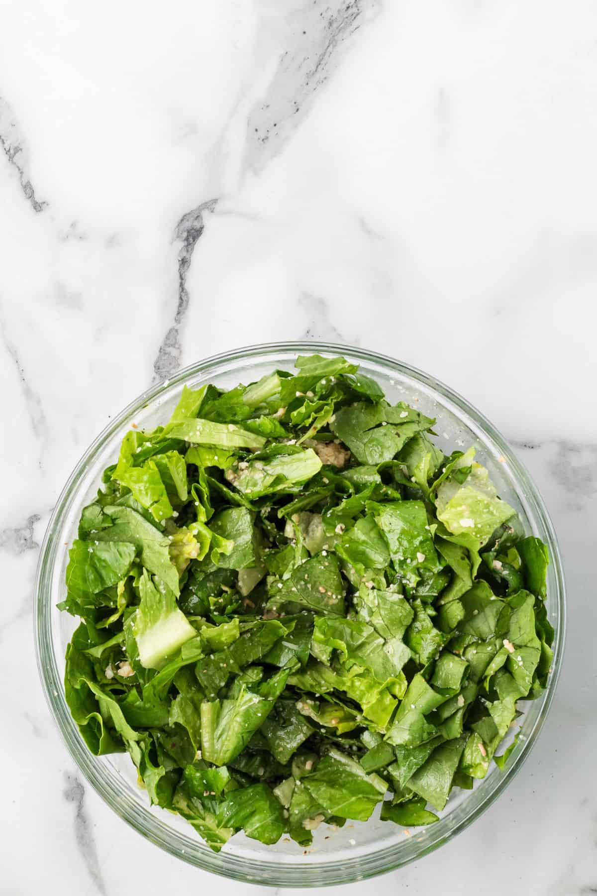 A glass bowl filled with chopped green leafy salad, placed on a white marble surface.