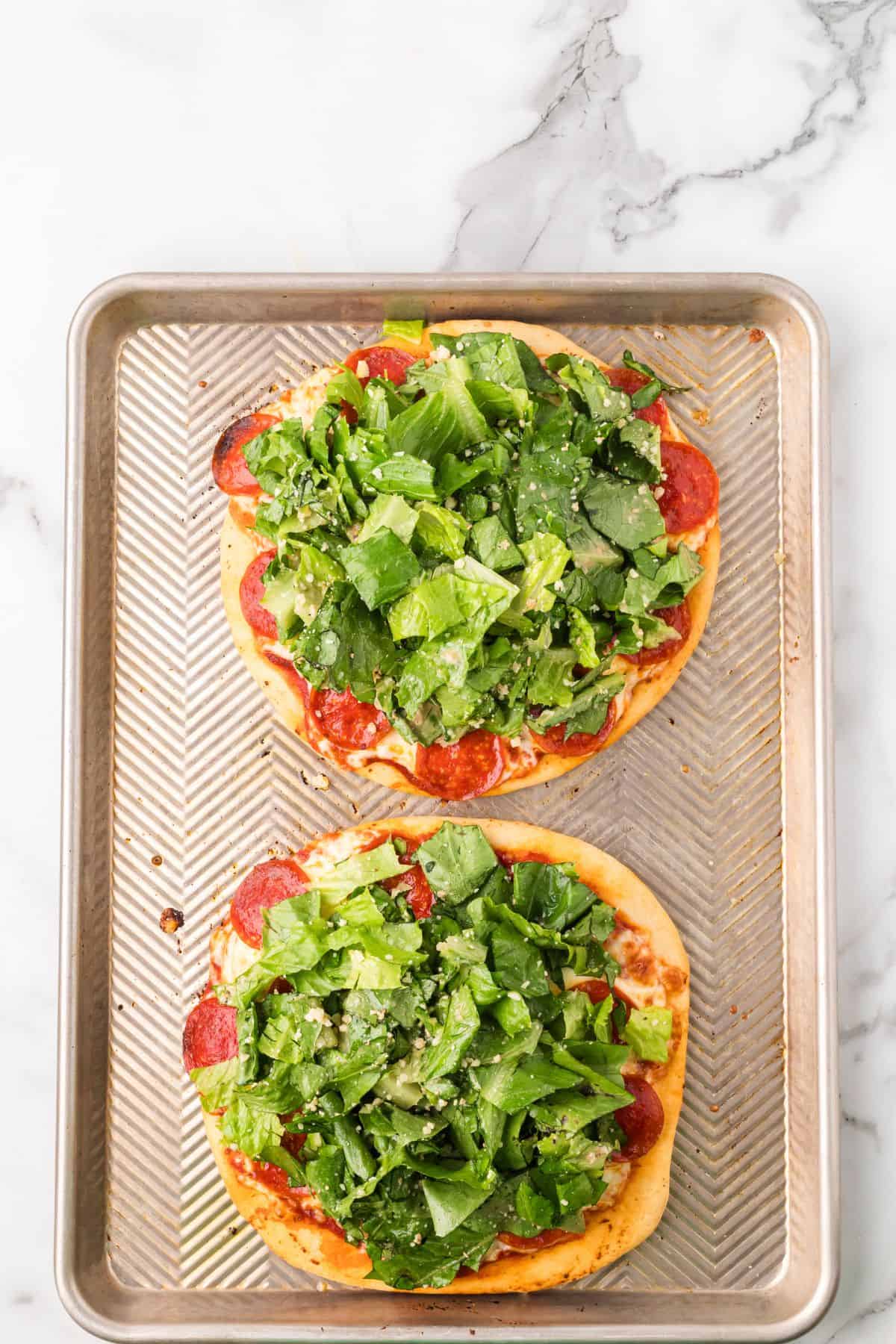 Two mini pepperoni pizzas topped with fresh green salad sit on a metal baking sheet on a marble surface.