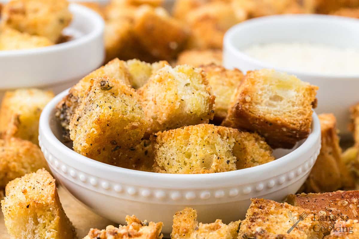 A white bowl filled with seasoned, golden brown croutons, with more croutons and a dip in the background.
