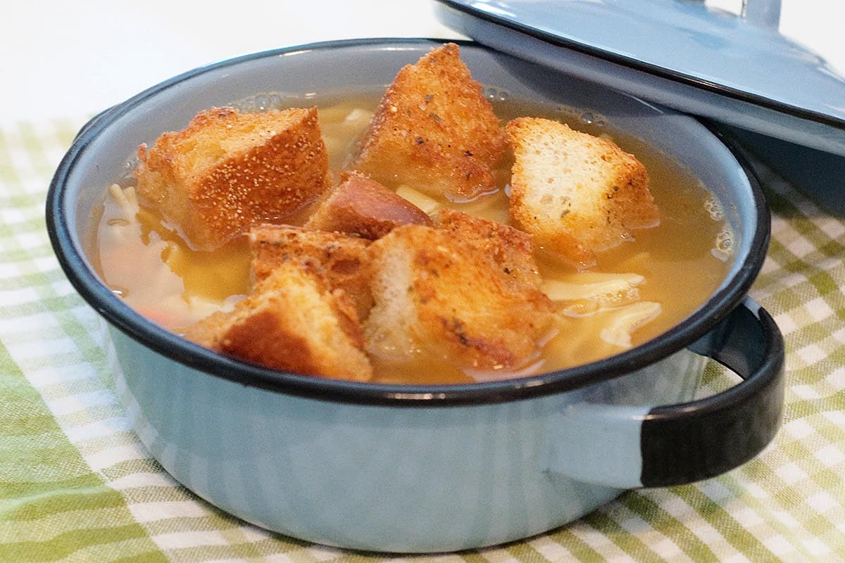 A blue pot of soup with noodles and croutons, sitting on a green and white checkered cloth.