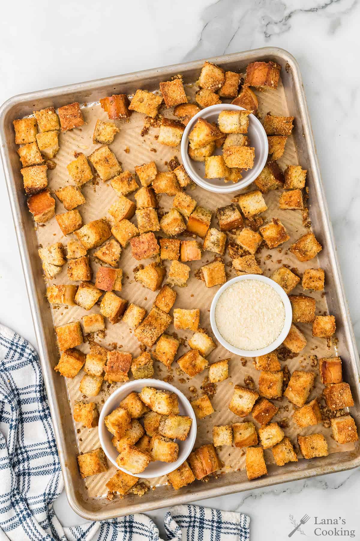 A baking sheet with seasoned croutons, bowls of croutons and parmesan cheese, and a blue plaid towel on the side.