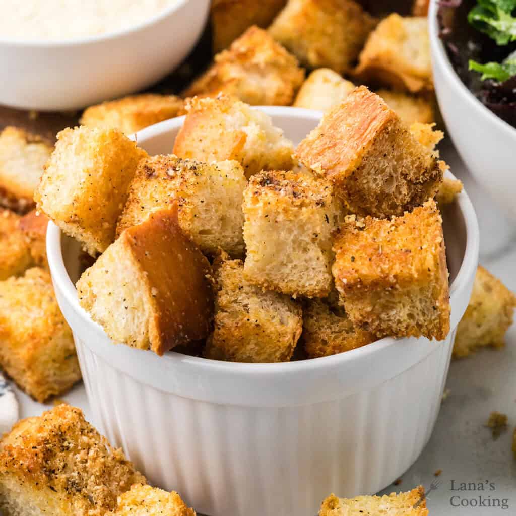 Crispy, seasoned croutons piled in a white ramekin with more scattered around on a table.