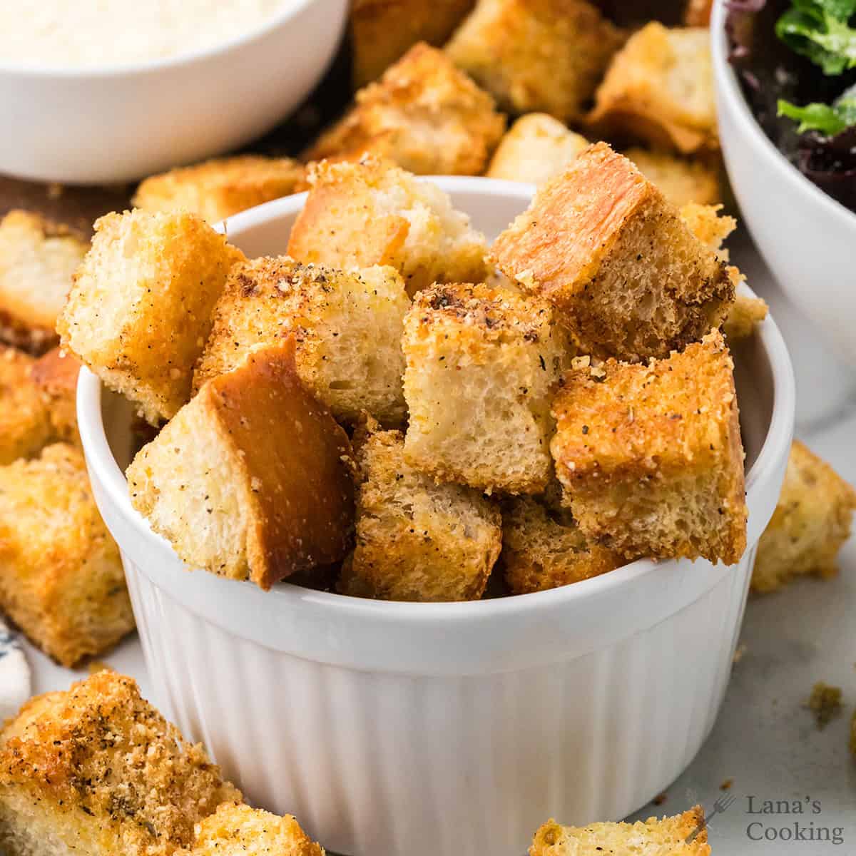 Crispy, seasoned croutons piled in a white ramekin with more scattered around on a table.