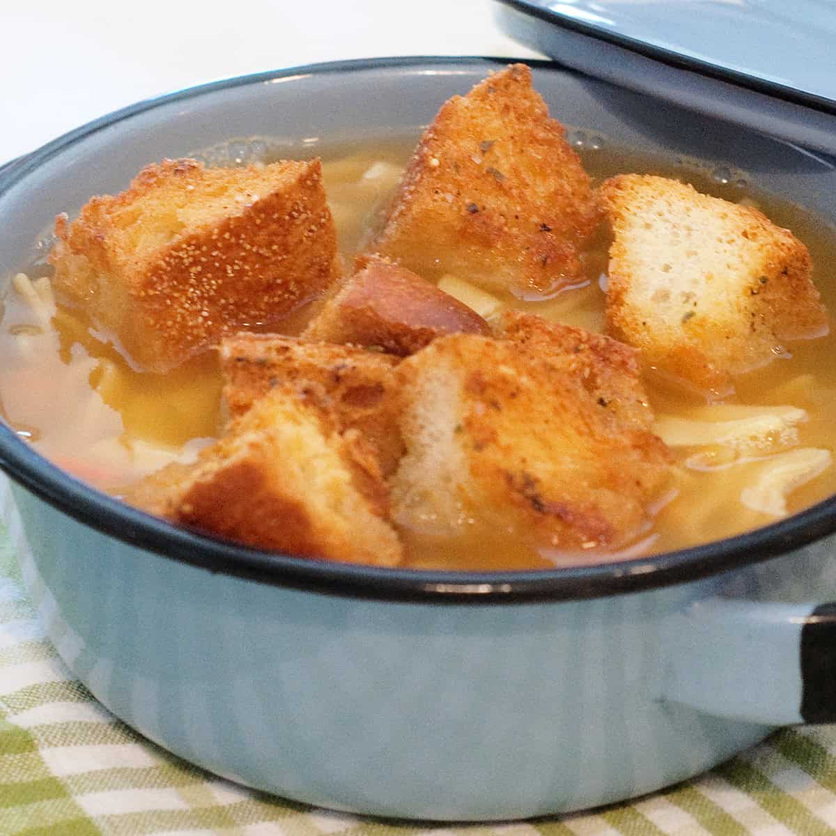 A blue bowl of soup with large croutons on top, sitting on a green and white checkered cloth.
