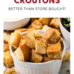 A white bowl filled with homemade croutons sits on a table with more croutons scattered around it.