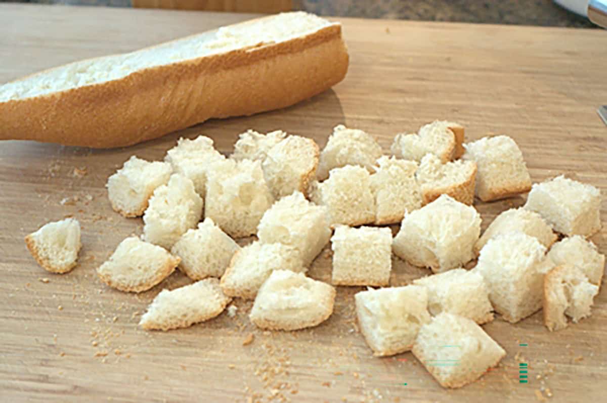 Cubed pieces of bread on a wooden cutting board with a partially sliced baguette.