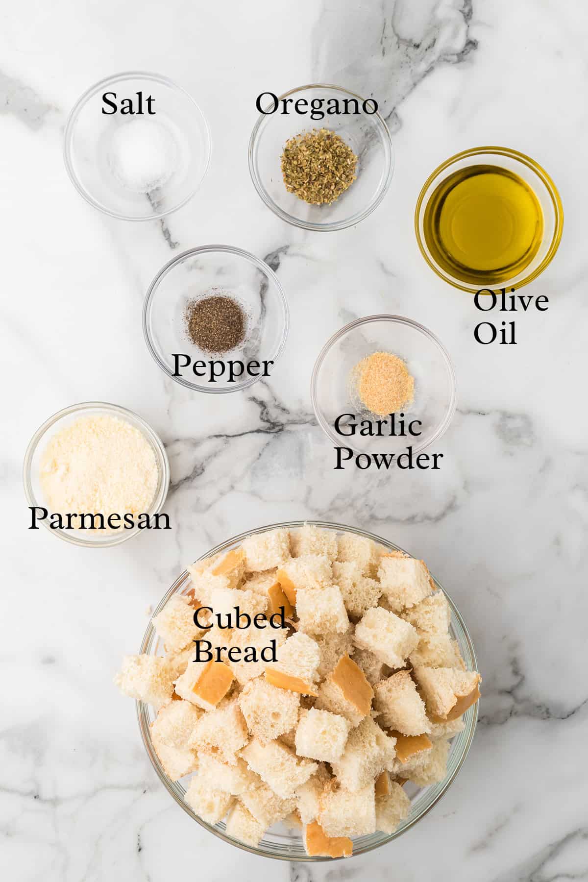 Bowls of cubed bread, parmesan, salt, pepper, oregano, garlic powder, and olive oil on a marble surface.