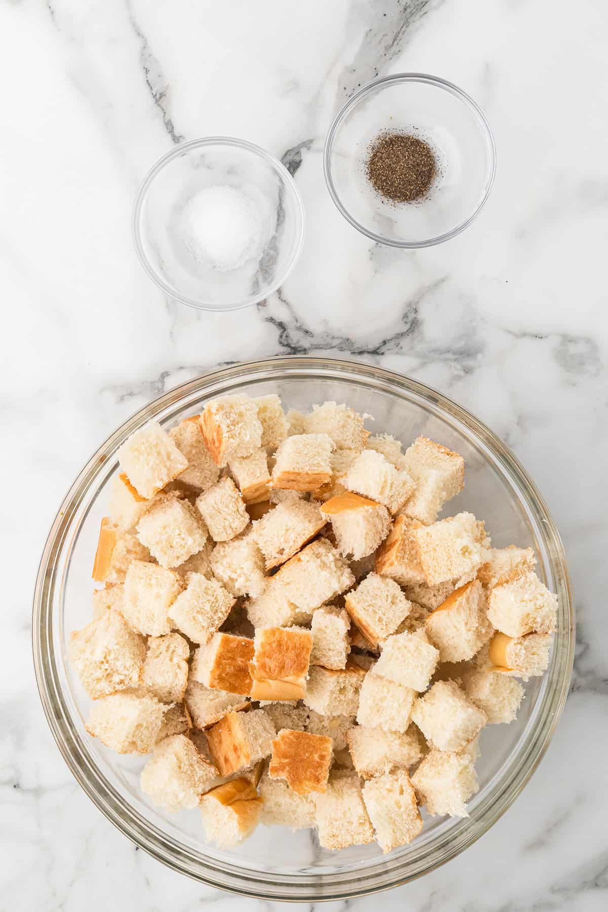 A glass bowl of cubed bread with two small bowls of salt and pepper on a marble countertop.