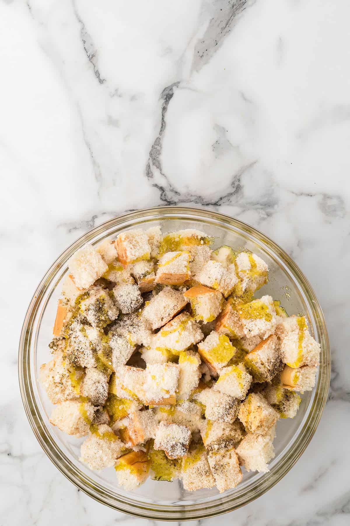 Cubed bread tossed with oil and seasonings in a glass bowl on a marble countertop.