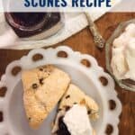 Two currant scones with jam and cream on a white plate, beside a jar of jam and bowl of whipped cream.