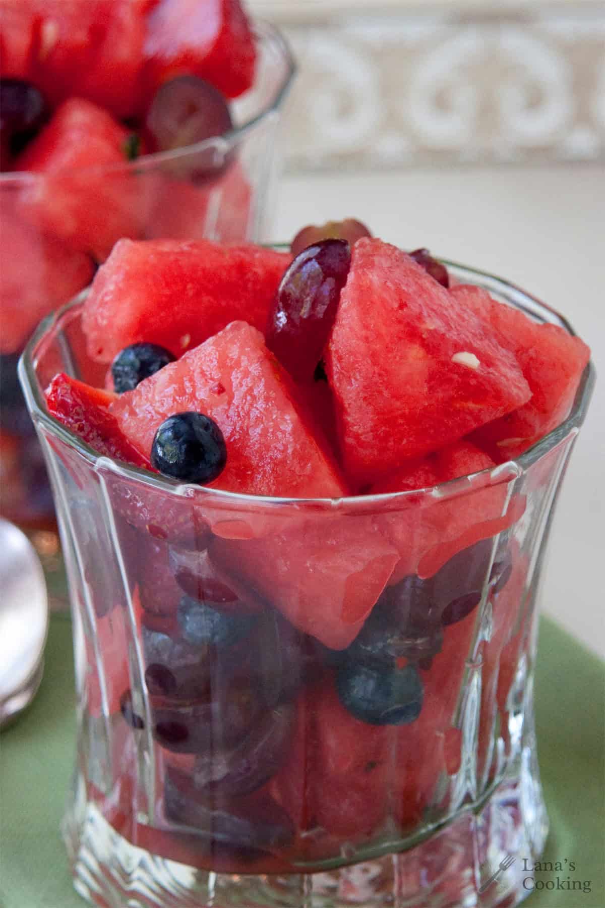 Glass filled with a refreshing watermelon salad featuring juicy watermelon chunks, blueberries, strawberries, and grapes with a ginger-mint syrup on a light green napkin.