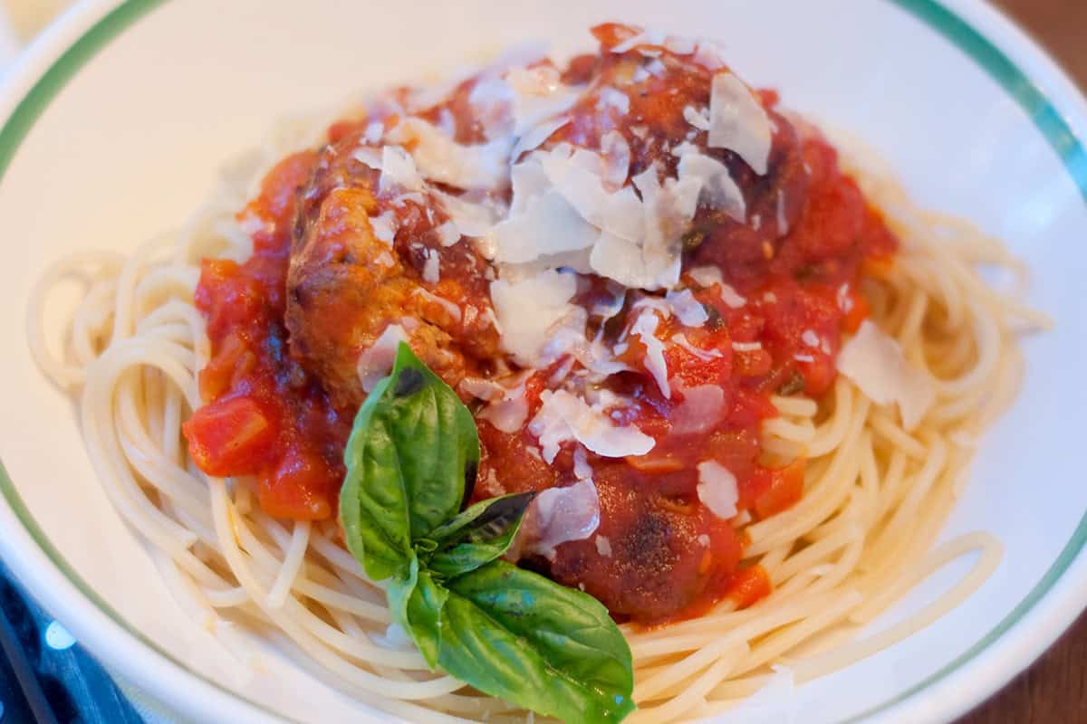 A bowl of spaghetti topped with tomato sauce, meatballs, shaved parmesan cheese, and a sprig of fresh basil.