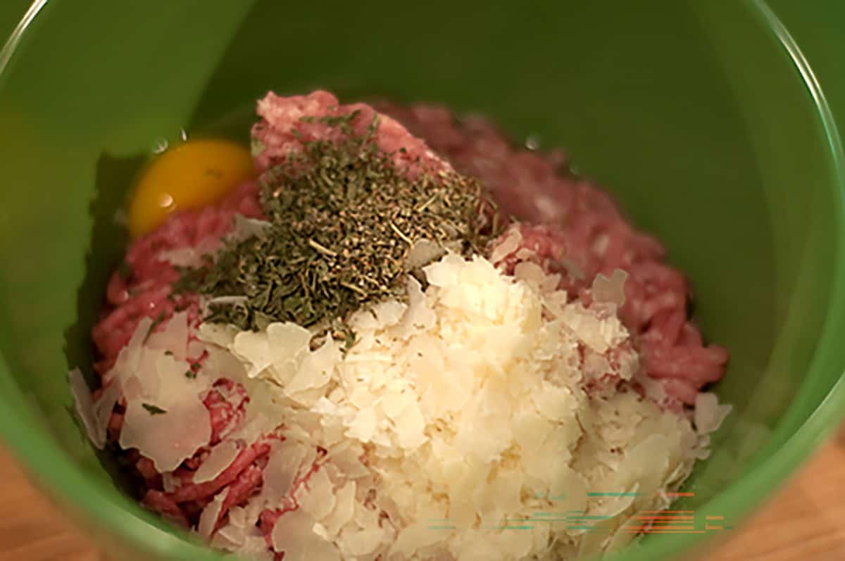 Raw ground meat, a cracked egg, chopped onions, and dried herbs are placed together in a green mixing bowl.