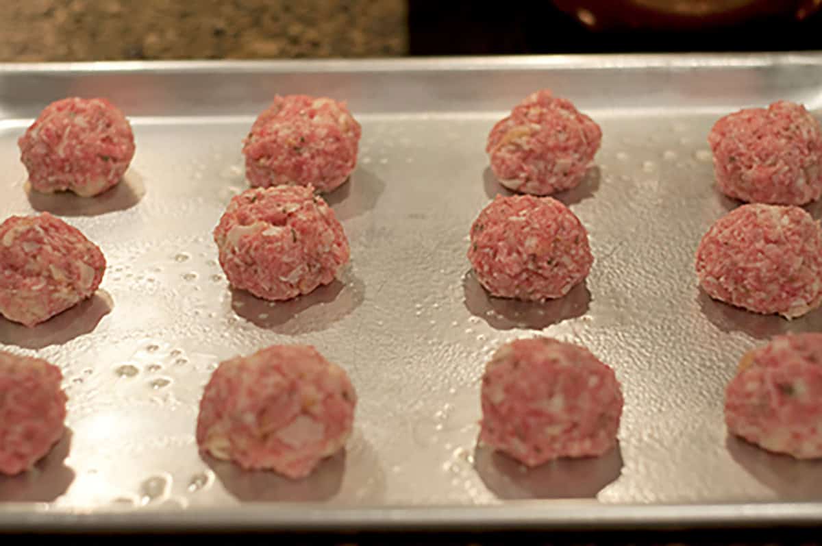 Raw meatballs are arranged in rows on a silver baking sheet, ready to be cooked. The tray is lightly oiled, and the meatballs appear to contain chopped onions or similar ingredients.