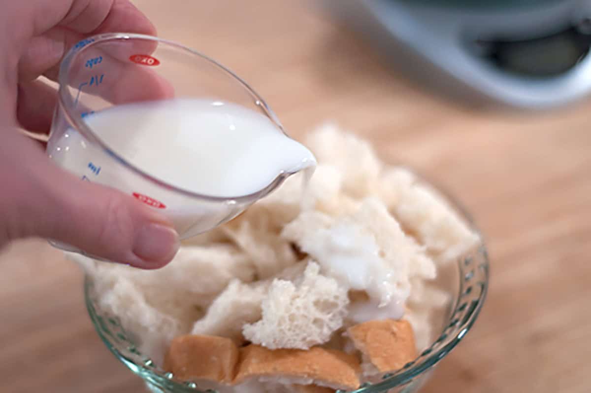 A hand pours milk from a small measuring cup onto pieces of white bread in a glass bowl, on a wooden surface.