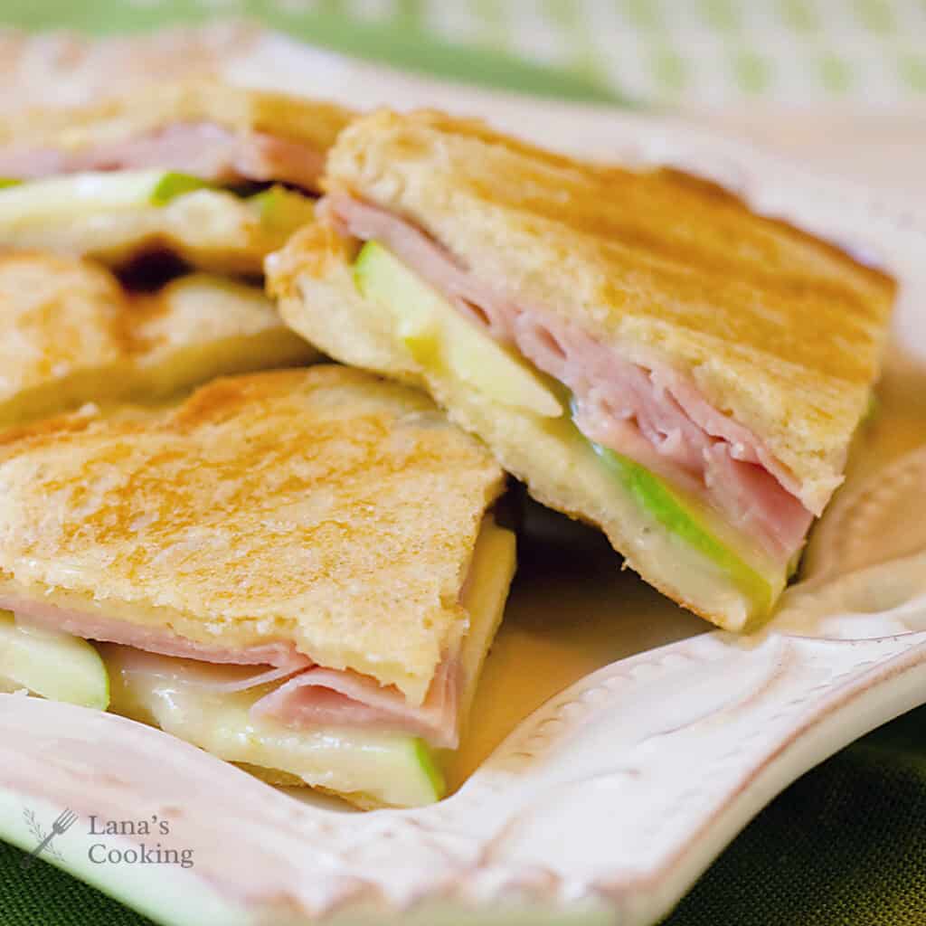 Three sandwich wedges filled with ham, cheese, and thin slices of green apple are stacked on a decorative white plate. The sandwiches are lightly toasted, and some melted cheese is visible.