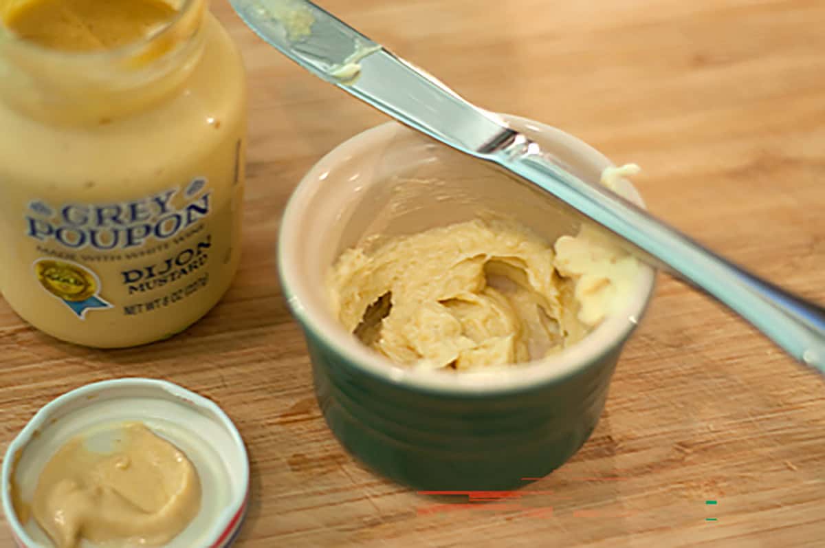 A jar of Dijon mustard, a small dish of mustard mixture, and a knife on a wooden surface.