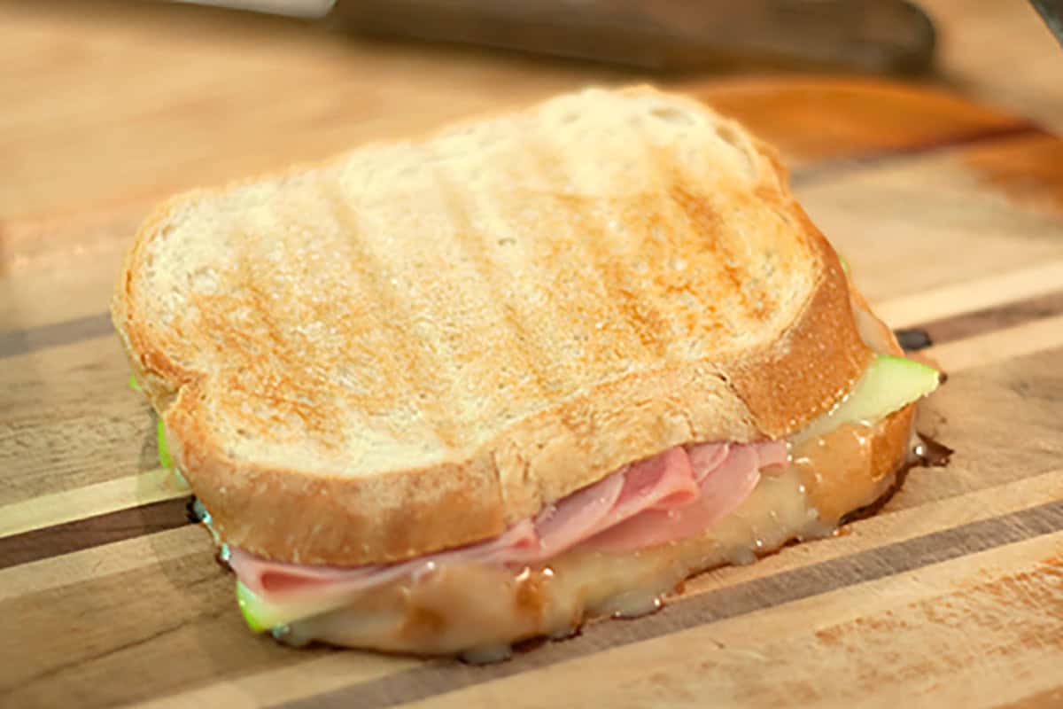 A grilled ham, apple, and brie panini with perfectly melted cheese on a wooden cutting board.