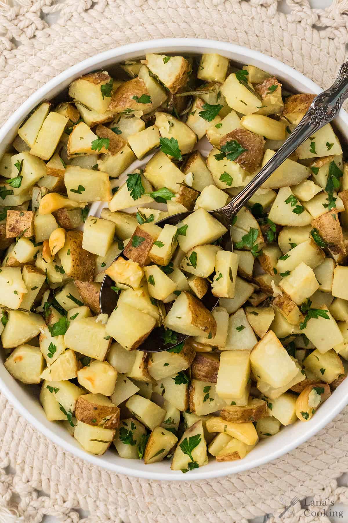 A white bowl filled with roasted potato cubes garnished with chopped parsley and garlic, with a serving spoon inside the bowl.