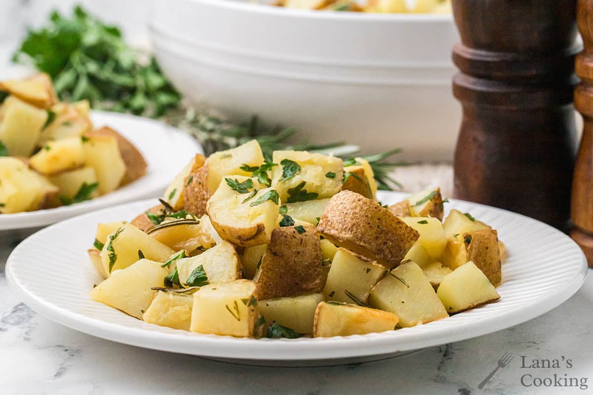 A plate of roasted potato cubes garnished with fresh herbs, with a bowl of more potatoes and pepper mills in the background.