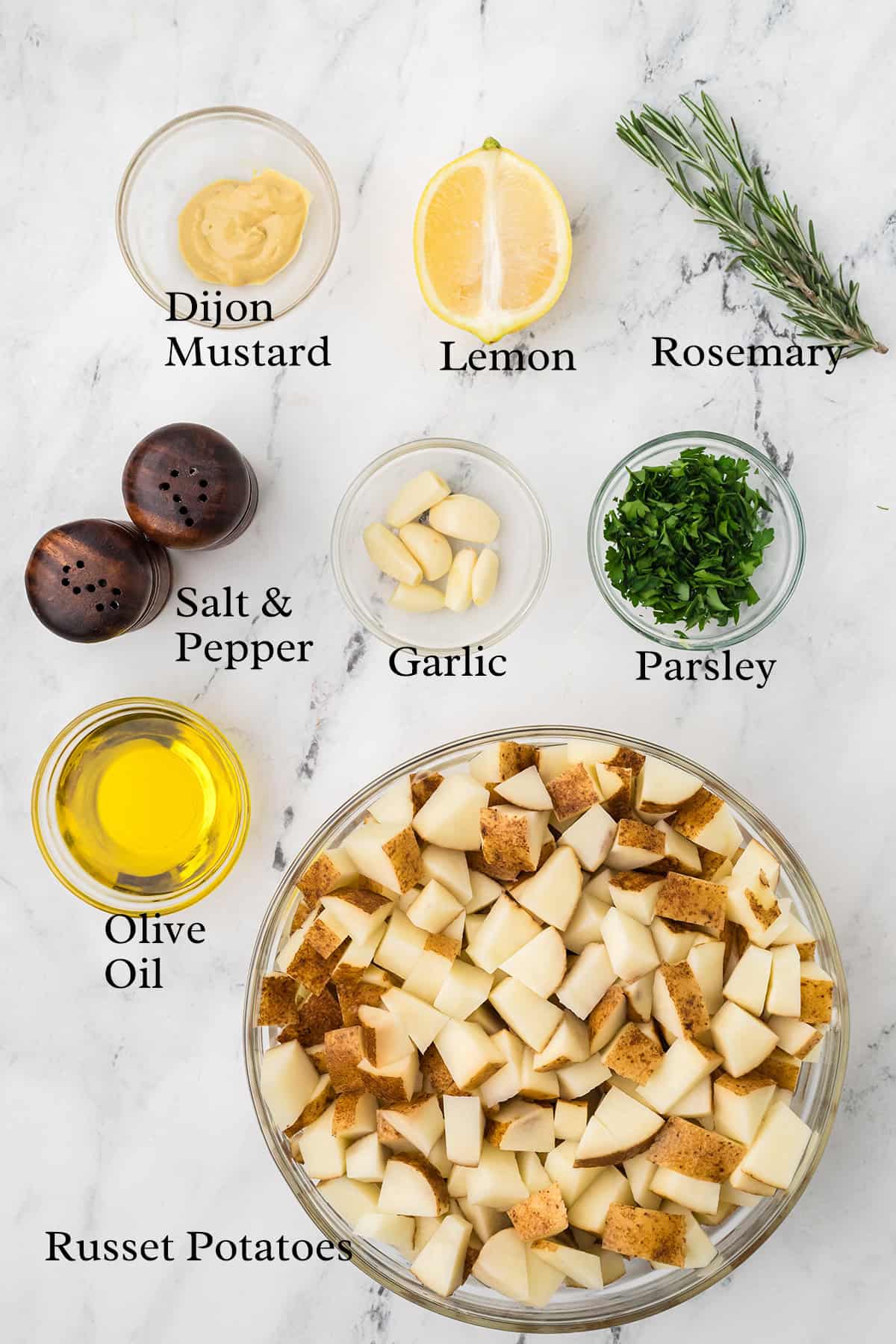 A marble surface displays bowls of chopped russet potatoes, olive oil, chopped parsley, garlic cloves, a lemon half, a sprig of rosemary, Dijon mustard, salt, and pepper.