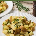 A plate of roasted potato salad garnished with chopped herbs sits on a white table, with another plate and fresh rosemary in the background.