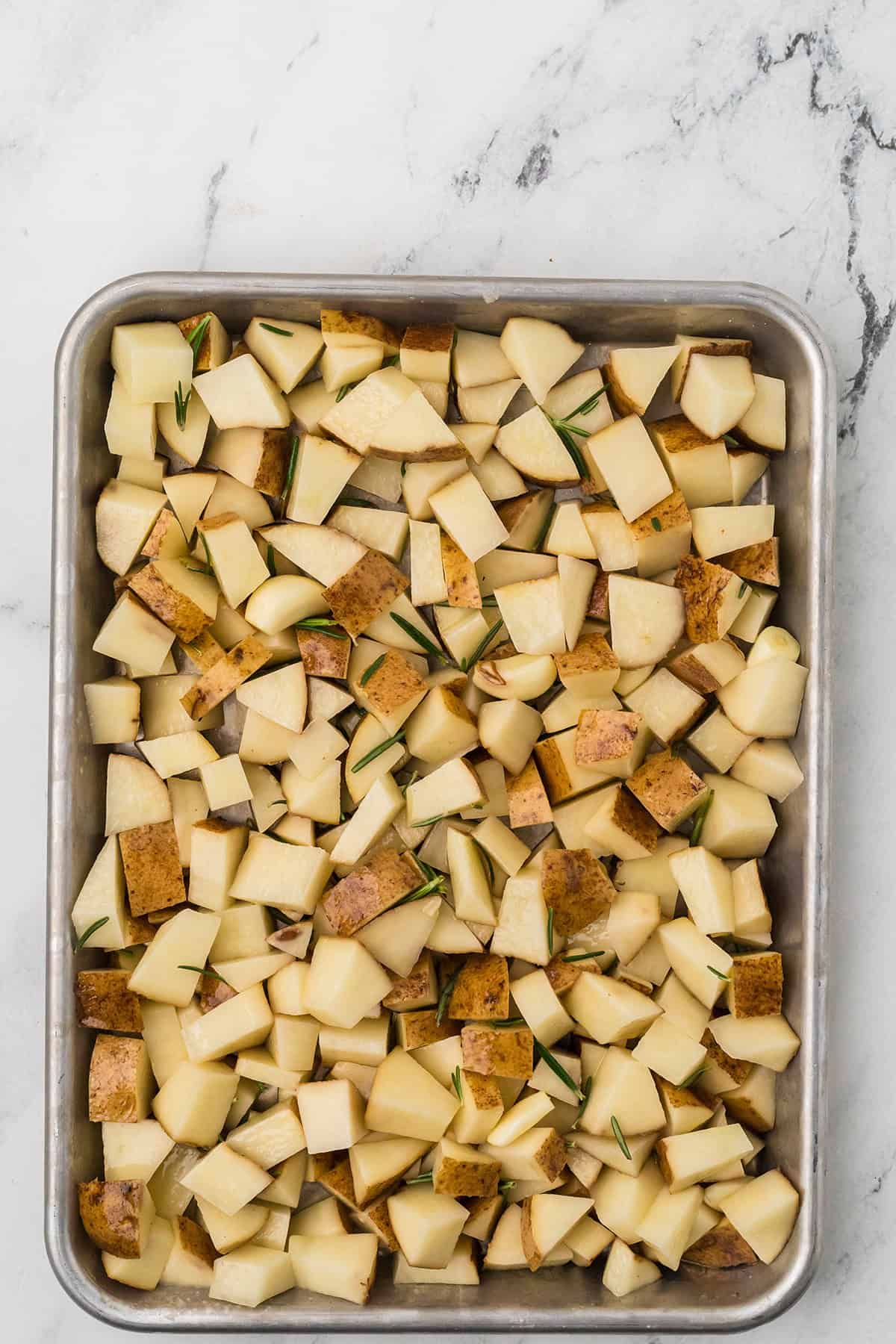 Cubed potatoes mixed with garlic, rosemary, and olive oil on a large baking sheet.