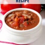 A bowl of hearty cowgirl beans with tomatoes and onions, beside fresh tomatoes and a red-striped napkin.
