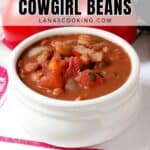 A white bowl filled with beans, chopped tomatoes, and onions sits on a white plate with a red-striped napkin. Large text above reads, Hearty Delicious Cowgirl Beans, with a website link below.