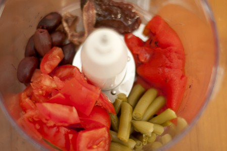 Food processor bowl containing olives, tomatoes, red bell peppers, anchovy, and green beans.