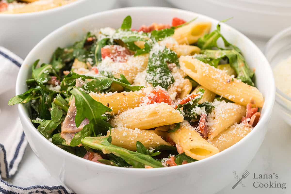 A bowl of penne pasta with arugula, tomatoes, and grated cheese on top.