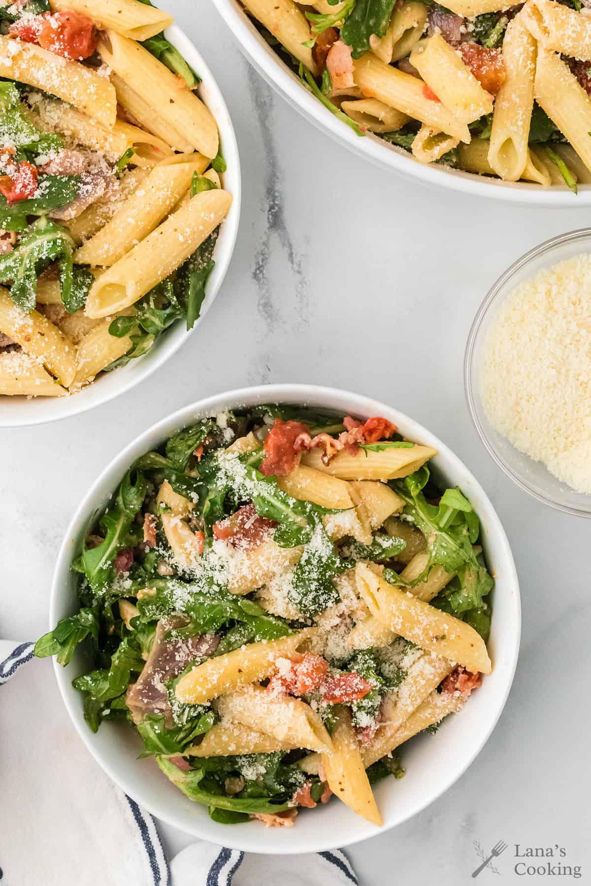 Three bowls of penne pasta with greens, tomatoes, and grated cheese on a white surface.