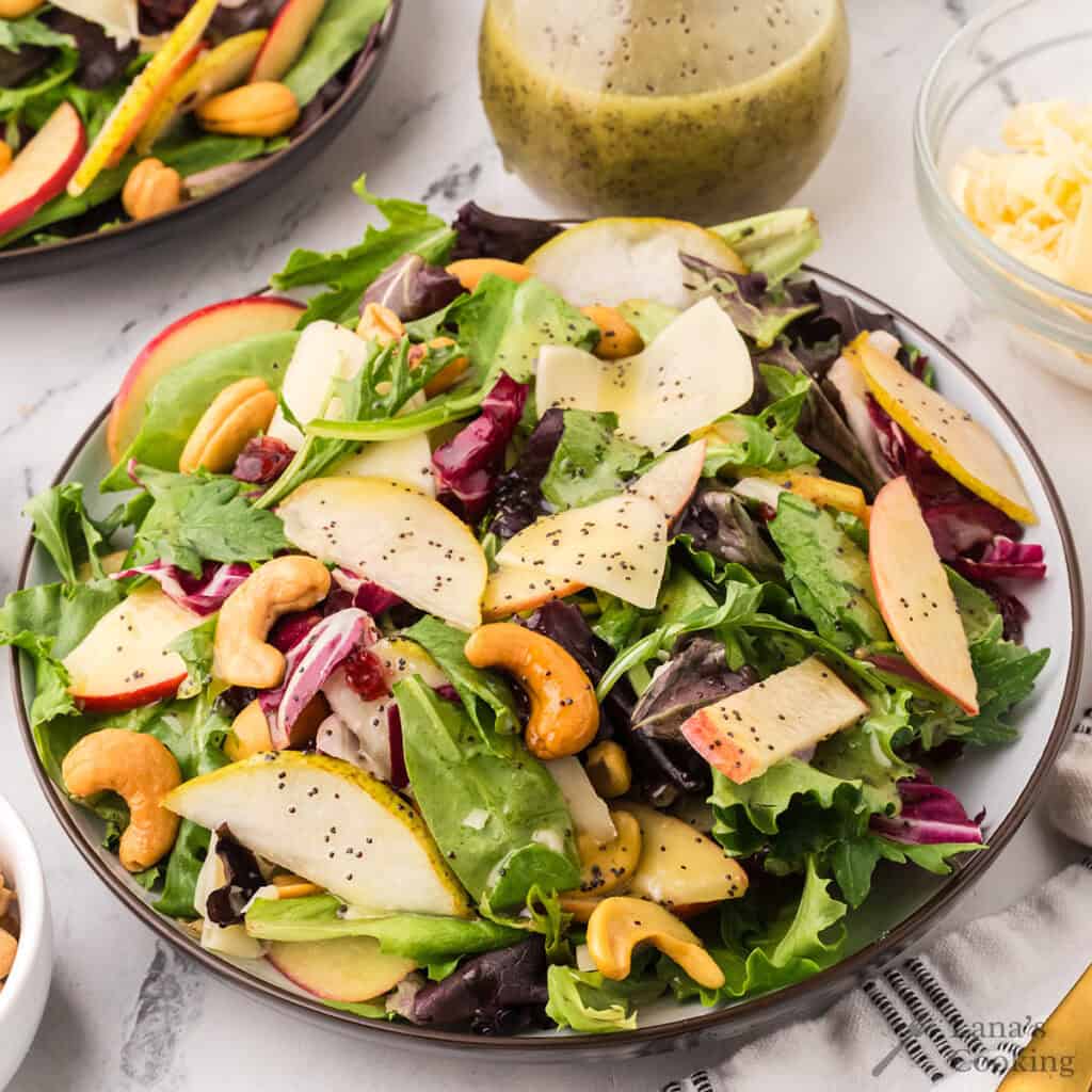 A plate of mixed green salad with sliced apples, pears, cashews, and shredded cheese, topped with poppy seed dressing. A small bowl of grated cheese and a jar of salad dressing are in the background.
