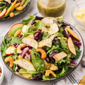 A plate of mixed green salad with sliced apples, pears, cashews, and shredded cheese, topped with poppy seed dressing. A small bowl of grated cheese and a jar of salad dressing are in the background.