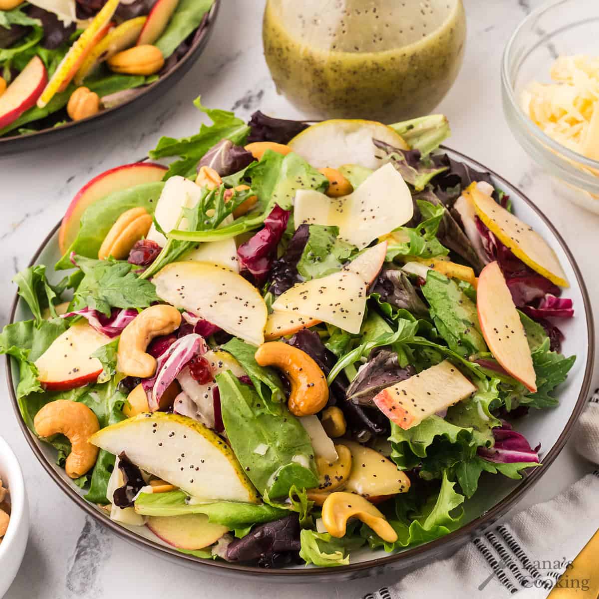 A plate of mixed green salad with sliced apples, pears, cashews, and shredded cheese, topped with poppy seed dressing. A small bowl of grated cheese and a jar of salad dressing are in the background.