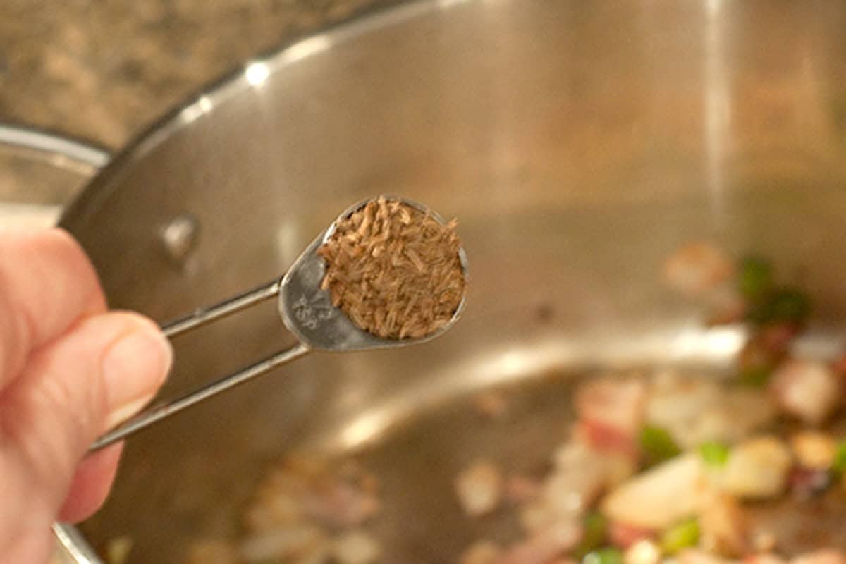 Add Caraway Seeds to Braised Cabbage A hand holds a teaspoon of caraway seeds over a pot of chopped onions and green peppers.
