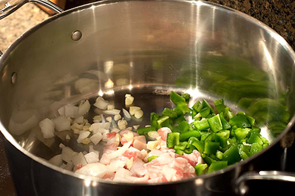 Starting Braised Cabbage Chopped onions, garlic, green bell peppers, and bacon in a stainless steel pot on a stovetop.