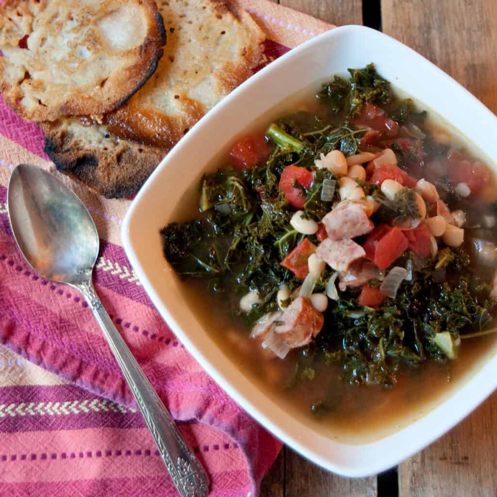 A bowl of soup with greens, beans, and sausage beside a spoon, bread, and a pink patterned napkin.