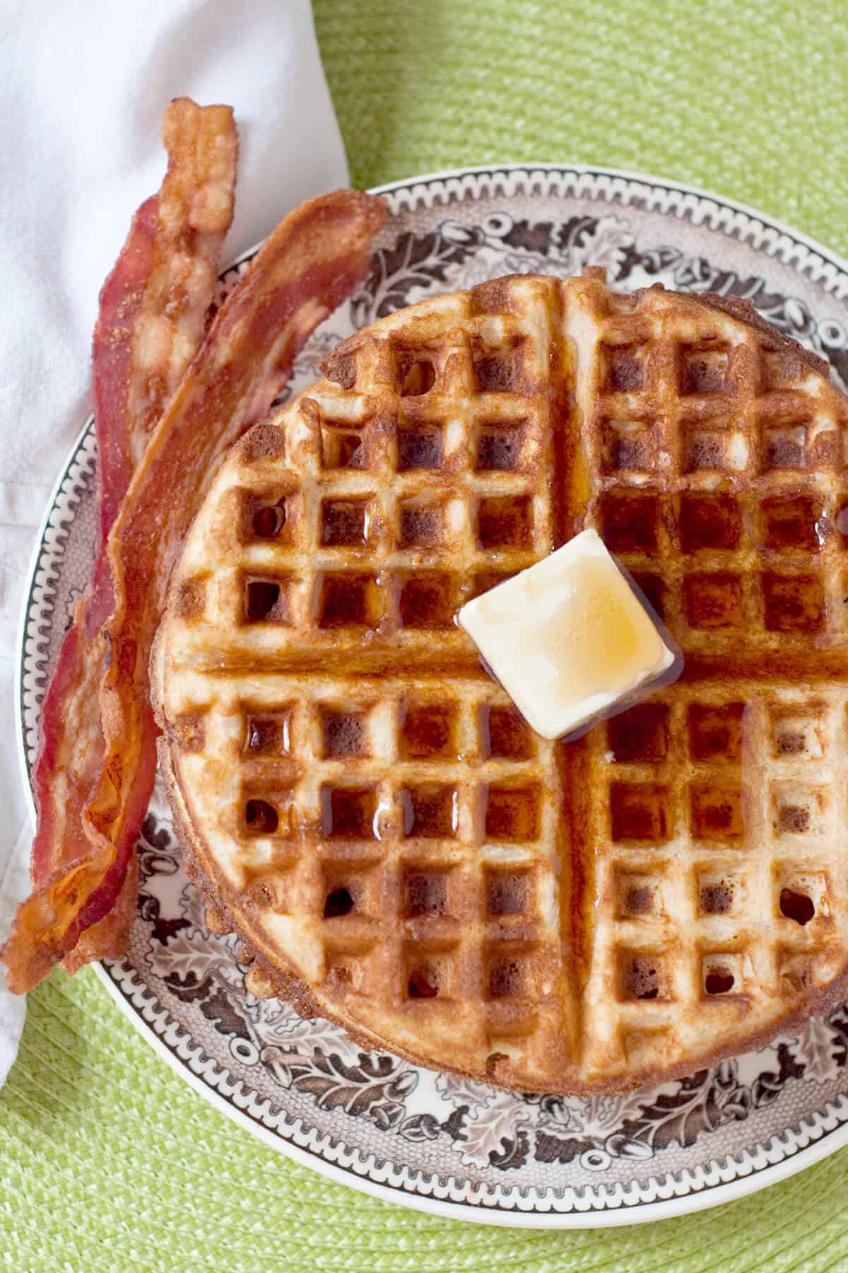 A stack of waffles with butter and syrup on top, bacon on the side.