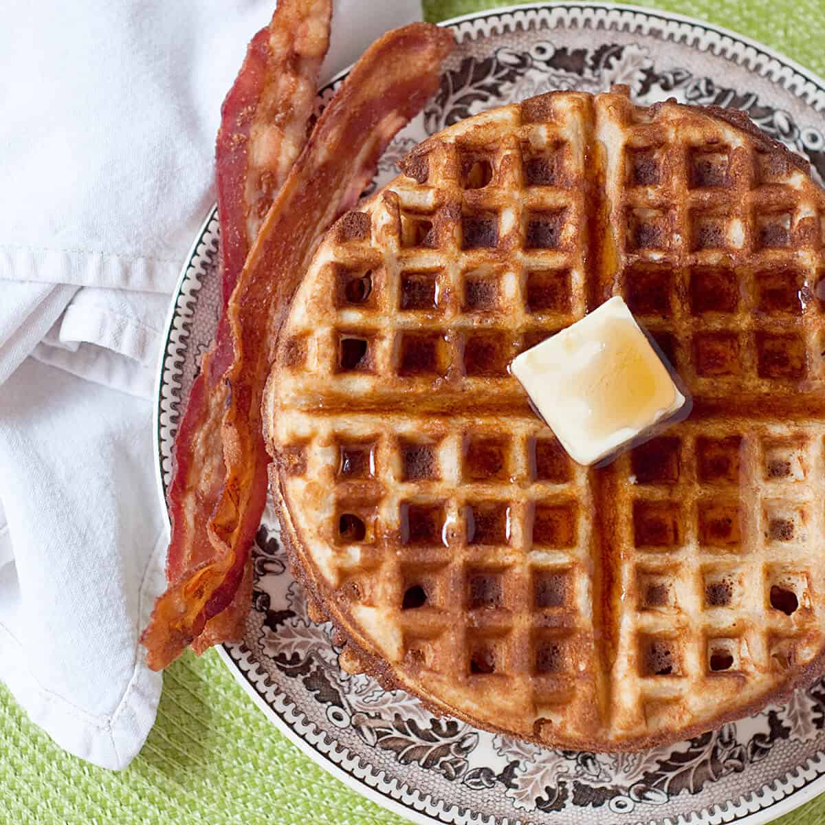 A stack of waffles with butter and syrup on top, bacon on the side.