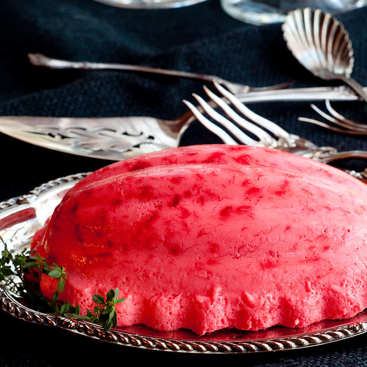 Raspberry jello mold on a silver serving tray with vintage serving pieces in the background.