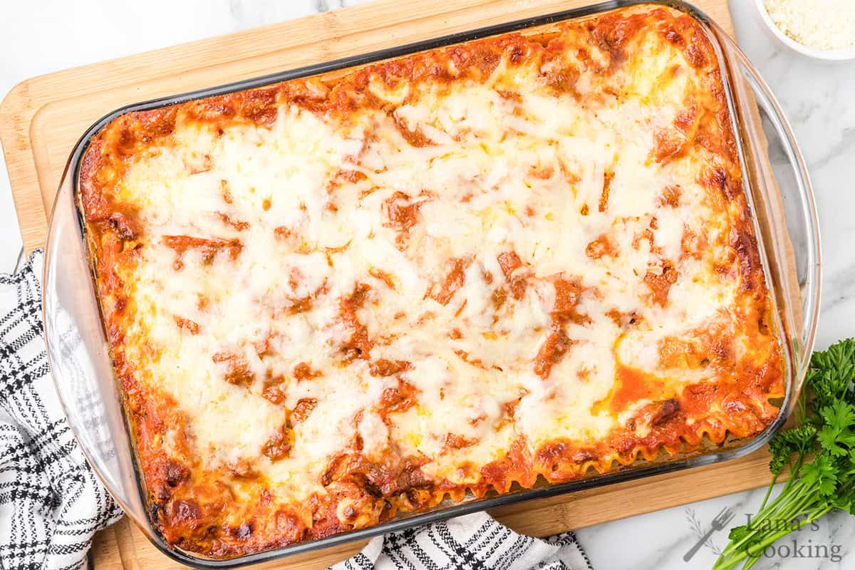 A baked lasagna in a glass dish on a wooden board, topped with melted cheese and tomato sauce.