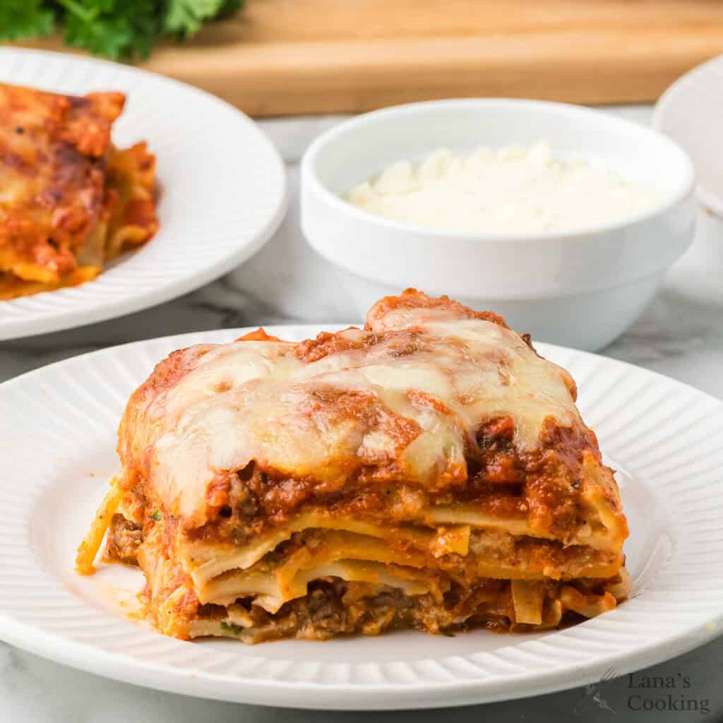 A slice of cheesy baked lasagna on a white plate with a bowl of grated cheese in the background.