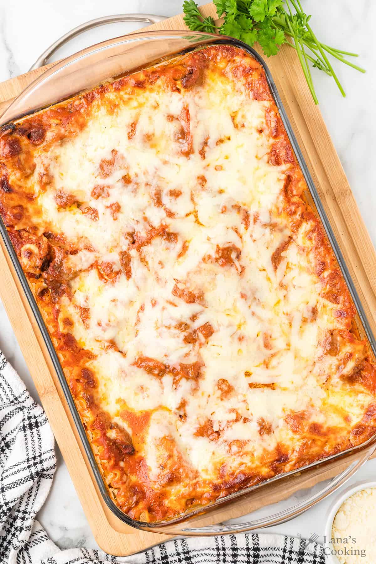 A glass baking dish filled with baked cheesy lasagna on a wooden board, with parsley and a towel nearby.