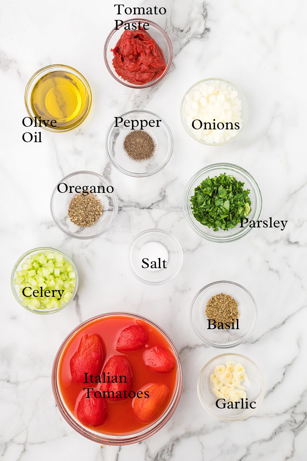 Ingredients for marinara sauce arranged on a marble surface, each labeled: tomatoes, herbs, oil, and seasonings.