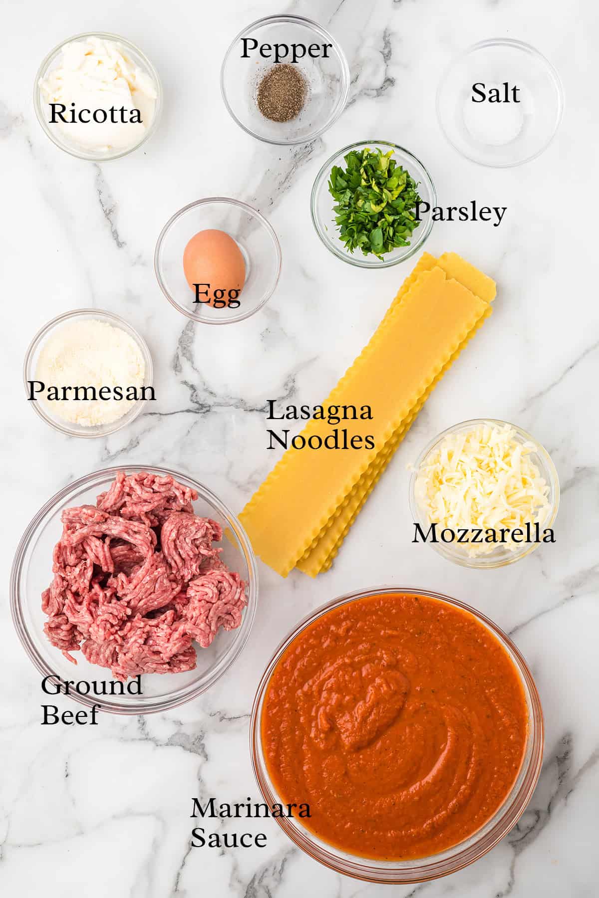 Lasagna ingredients on a marble surface: ricotta, parmesan, ground beef, egg, noodles, sauce, cheese, parsley, salt, pepper.