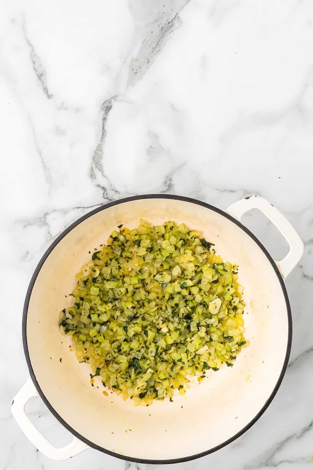 Overhead view of chopped celery and onions sautéing in a white pot on a marble countertop.