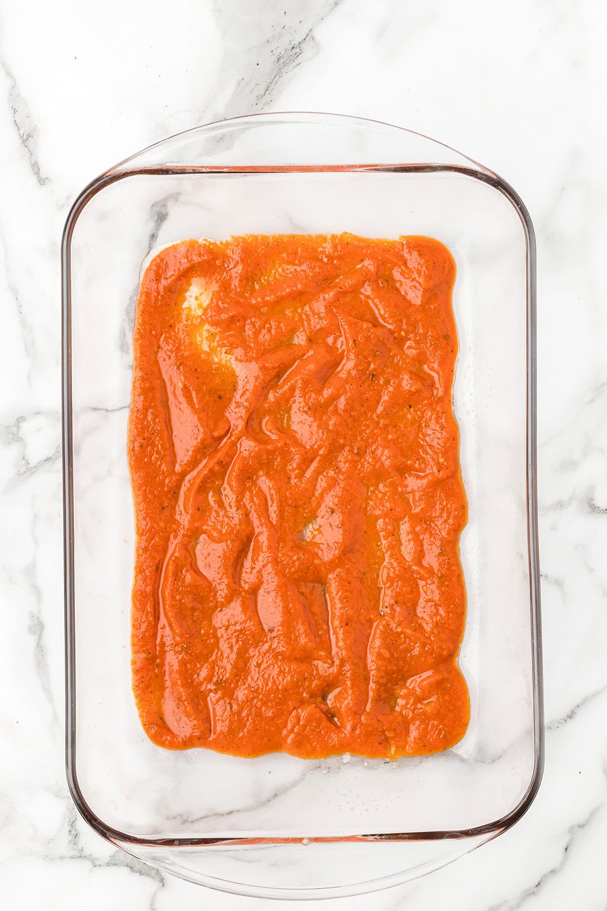 A glass baking dish with a layer of red marinara sauce on a white marble surface.